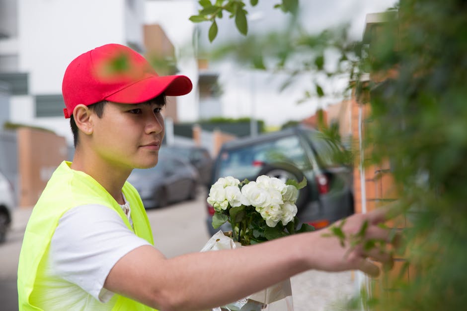 A young male florist wearing a red cap and a neon yellow vest stands outdoors during daytime, delivering a floral gift consisting of fresh white hydrangeas arranged in a small bouquet. He is holding t