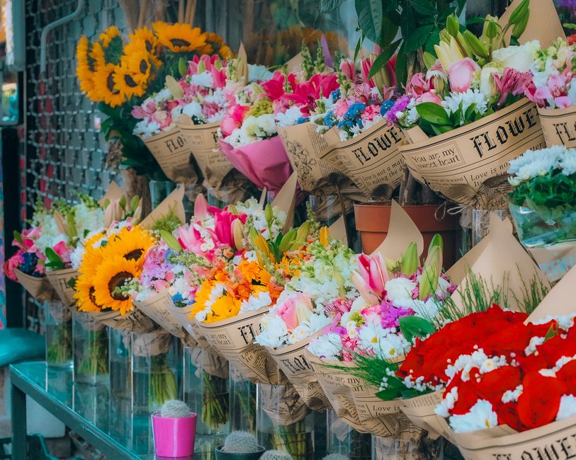 A vibrant display of freshly arranged flower bouquets wrapped in rustic newspaper, featuring a variety of blooms including bright yellow sunflowers, pink tulips, white lilies, orange marigolds, and re