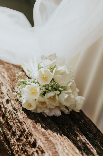 A delicate bridal bouquet featuring soft white flowers, including freesias and baby's breath, arranged with lush green foliage. The bouquet rests on a rustic wooden surface with a textured, weathered 