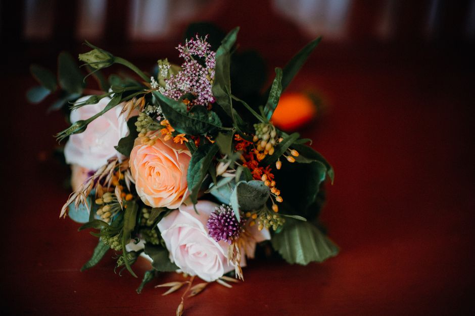 A close-up view of a floral bouquet featuring soft peach and pale pink roses, accented with small purple, orange, and white flowers, along with lush green foliage. The bouquet is arranged in a natural
